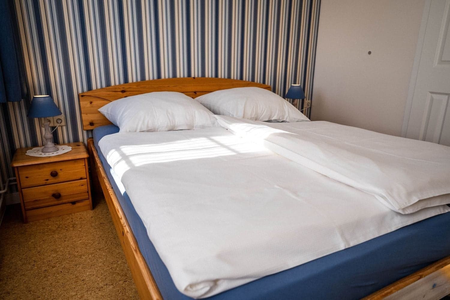 Doppelbett mit hellem Holzgestell, zwei Nachttischen mit blauen Lampen und blau gestreifter Tapete