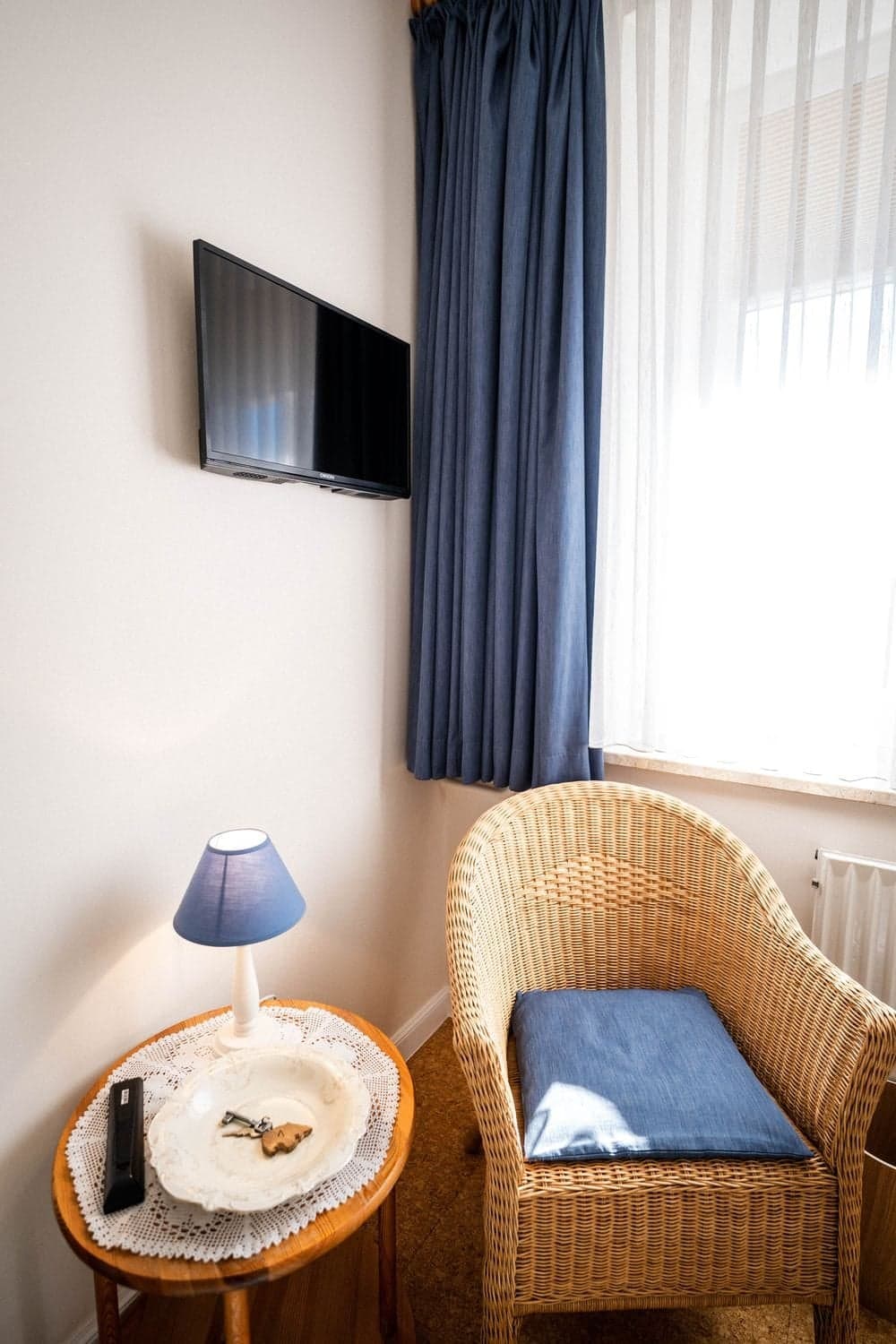 Zimmerecke mit Fernseher an der Wand, blauem Vorhang, Korbsessel und Beistelltisch mit Schlüssel auf einem Teller