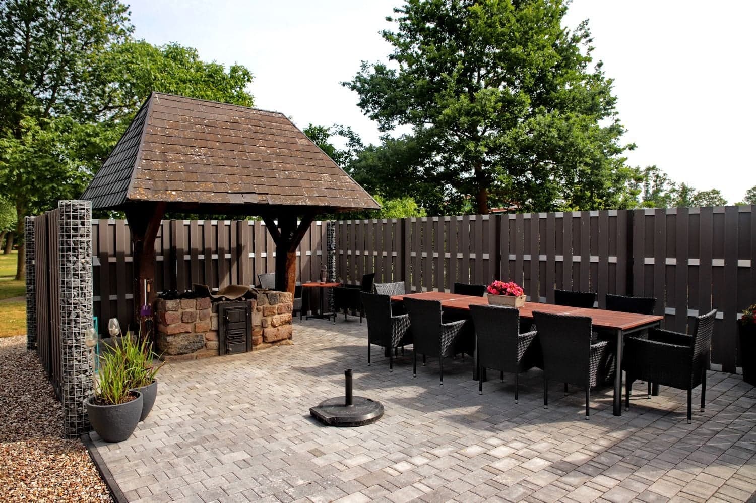 Gepflasterte Terrasse mit überdachtem Grillplatz, großem Holztisch und schwarzen Stühlen vor einem Holzzaun