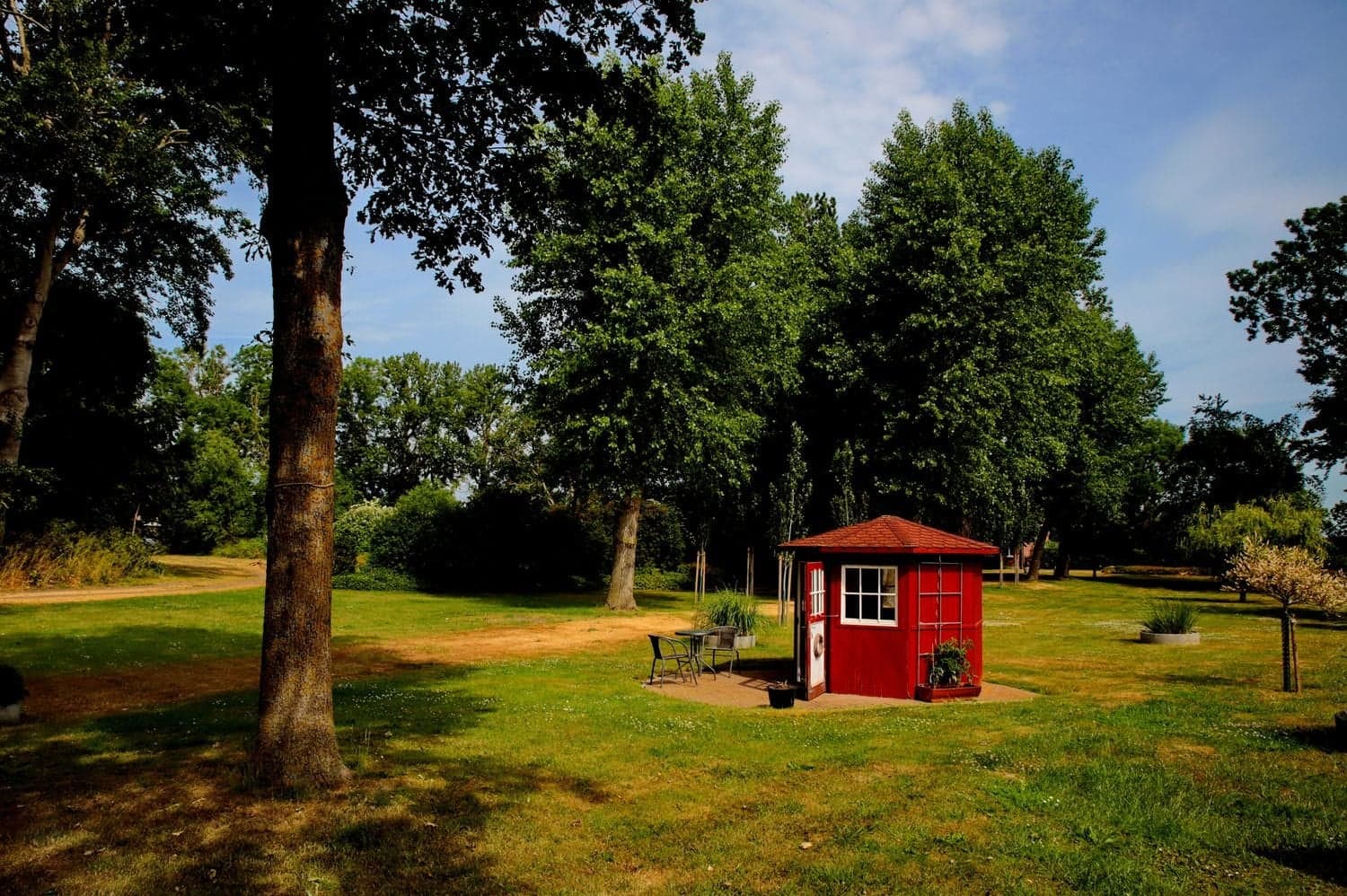 Rotes Gartenhäuschen mit Sitzplatz auf einer Wiese zwischen hohen Bäumen